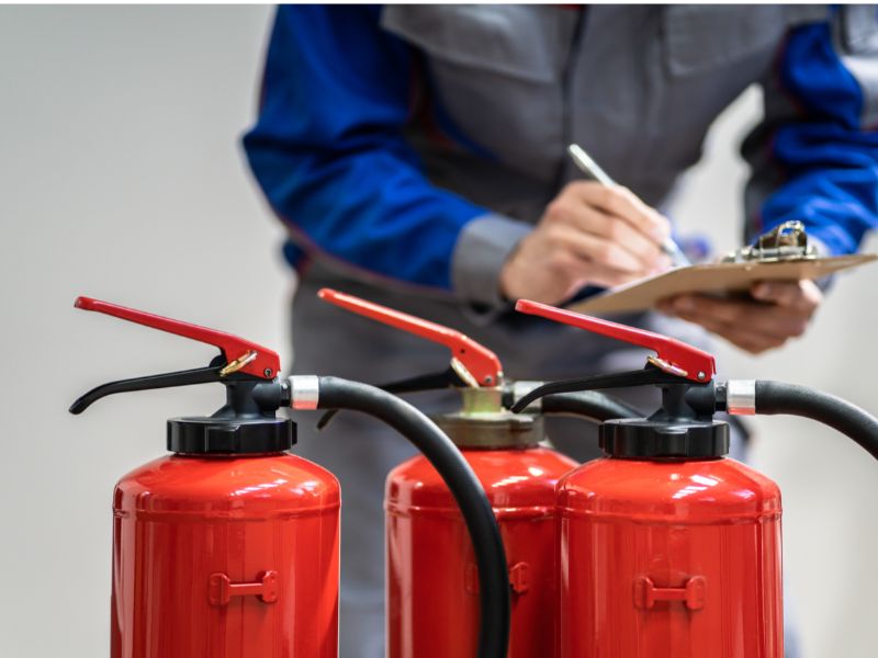 На передньому плані знаходяться два вогнегасника, на задньому плані чоловік в синьому комбінезоні із планшетом у руках робить записи
