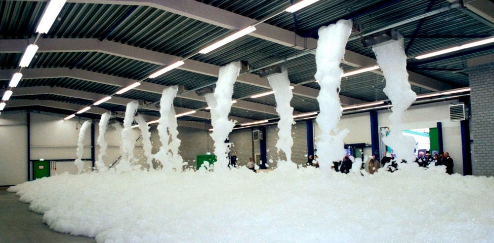 Велике складське приміщення із металевою кришею і білими лампами по періметру всього приміщення. В центрі знаходиться установка із якої надходить розчин із піною для гасіння. Також у лівому кутку фото можна побачити групу людей у поліцейській формі що стоять біля вихода із цього ангару.