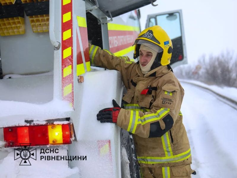 Рятувальник ДСНС Рівненщини у захисному шоломі та формі стоїть біля білого пожежно-рятувального автомобіля на засніженій трасі під час хуртовини