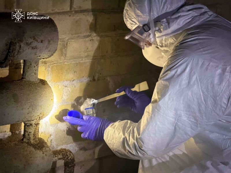 Специалист ГСЧС в белом защитном костюме и фиолетовых перчатках берет пробу вещества со стены в контейнер для химического анализа.