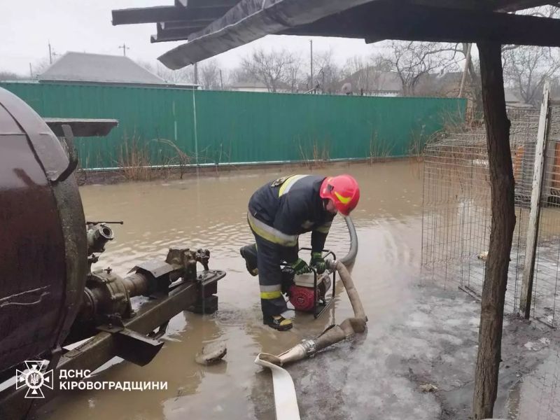 Спасатель ГСЧС в защитной экипировке запускает переносную мотопомпу для откачки воды с затопленного частного двора в Кировоградской области. На заднем плане зеленый забор и затопленная территория.