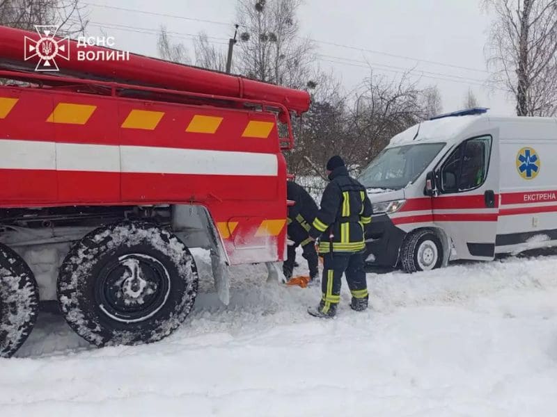 Пожарный автомобиль ГСЧС Волынской области буксирует машину скорой помощи, застрявшую в глубоком снегу на заснеженной сельской дороге.