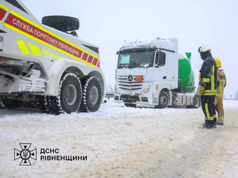 Тяжелая спецмашина ГСЧС Ровенской области с надписью «Служба спасения» на белом борту стоит перед белой фурой Mercedes-Benz на полностью заснеженной дороге во время зимней непогоды