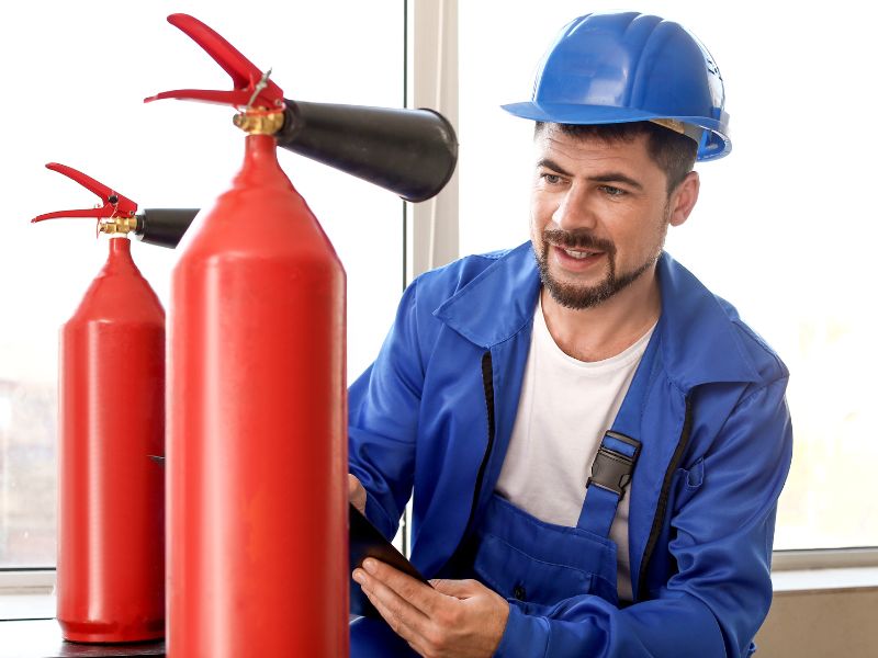 На фото чоловік в синьому робочому костюмі перевіряє стан вогнегасників, на ньому каска синього кольору та перед ним три вогнегасники червоного кольору